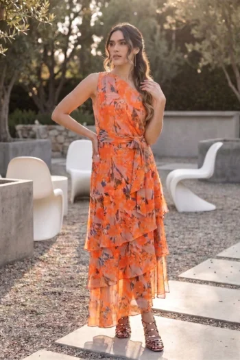 Woman in an orange floral maxi dress standing outdoors, hands on hip, sunlight filtering through trees.