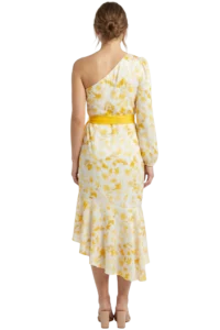 Back view of a woman wearing a white and yellow floral one-shoulder dress with a wide yellow sash belt, standing barefoot.