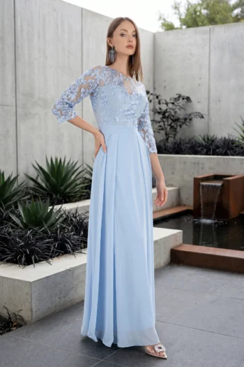 Woman wearing a light blue lace-shoulder gown stands outdoors by concrete walls, posing with one hand on her hip.