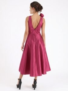 Back view of a woman wearing a magenta satin sleeveless tea-length dress with a deep V back, a waist seam, and a large bow detail at the left shoulder; she pairs it with black high-heeled sandals.