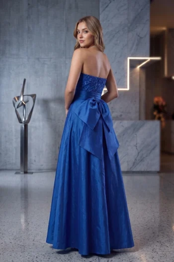 Back view of a woman in a royal blue strapless gown with a large bow at the waist, standing in a marble interior.] ,