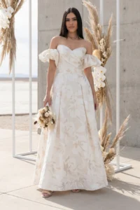 Woman in an off-shoulder, ivory floral gown standing outdoors among tall pampas grass arrangements.