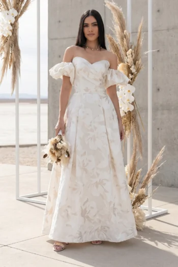 Woman in an off-shoulder, ivory floral gown standing outdoors among tall pampas grass arrangements.