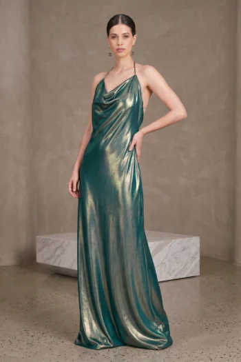 Model modeling a teal metallic halter gown with one hand on her hip against a neutral backdrop.