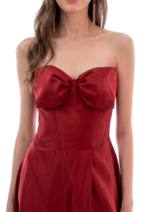 Woman wearing a red strapless satin dress with a large front bow and gold dangling earrings, single-portrait cutout against a dark background.