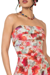 Woman in a strapless, multicolored floral dress wearing a gold statement necklace with colorful gems and red earrings; close focus on neckline and shoulders with a black background.