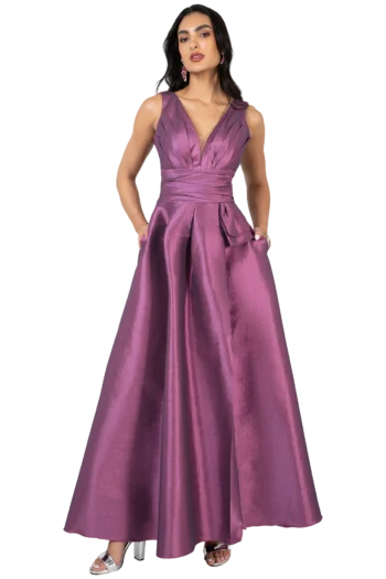 Woman in a shiny purple evening gown with a wrapped waist, hands in pockets, standing in metallic heels against a black background