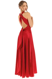 Woman in a red satin backless evening gown with crisscross straps, looking over her shoulder with dangling earrings—formal attire.