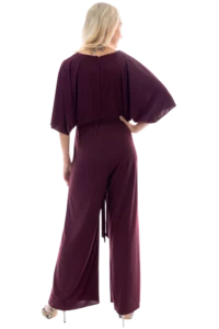 Back view of a person wearing a burgundy jumpsuit with wide sleeves, blonde hair, and a tattoo at the neck; hands on hips.