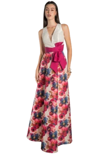 Woman wearing a white sleeveless V-neck top with a bright pink sash and a long floral skirt, posing against a black background.