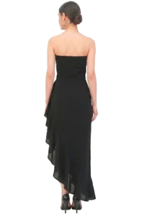 Back view of a woman in a strapless black evening gown with a layered, asymmetrical hem and high-heeled shoes, hair in a bun with large hoop earrings