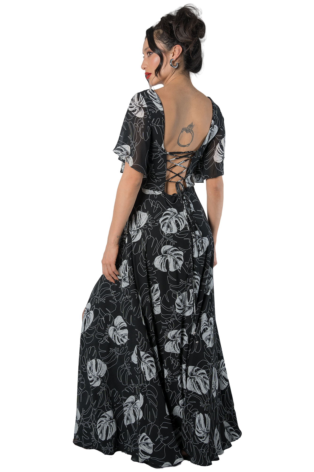 Woman in a black backless dress with white leaf pattern, facing left; lace-up back detail visible and a tattoo on her upper back.