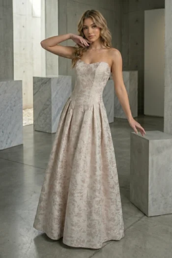 Woman in a pale pink strapless floral-embossed evening gown posing in a marble-and-concrete interior, one arm raised to her chin.