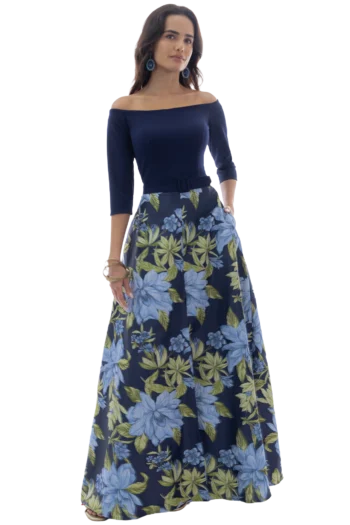 Woman in navy off-shoulder top and long blue floral skirt, with blue earrings, standing with one hand in her pocket.