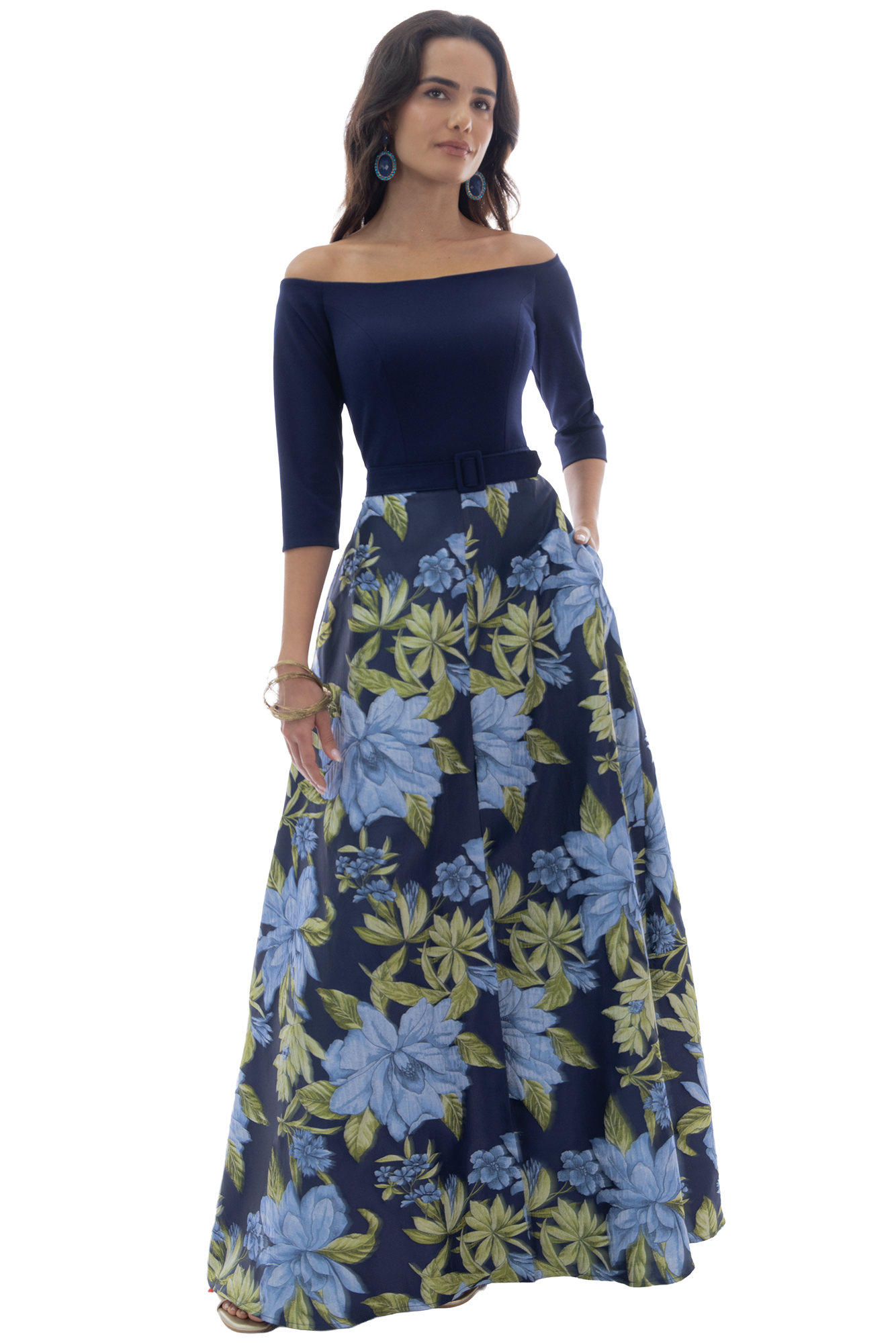 Woman in navy off-shoulder top and long blue floral skirt, with blue earrings, standing with one hand in her pocket.