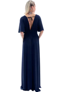 Woman seen from behind in a navy blue, floor-length dress with a deep V back and smocked waist; blonde hair with a clip and a tattoo at the neck/upper back.