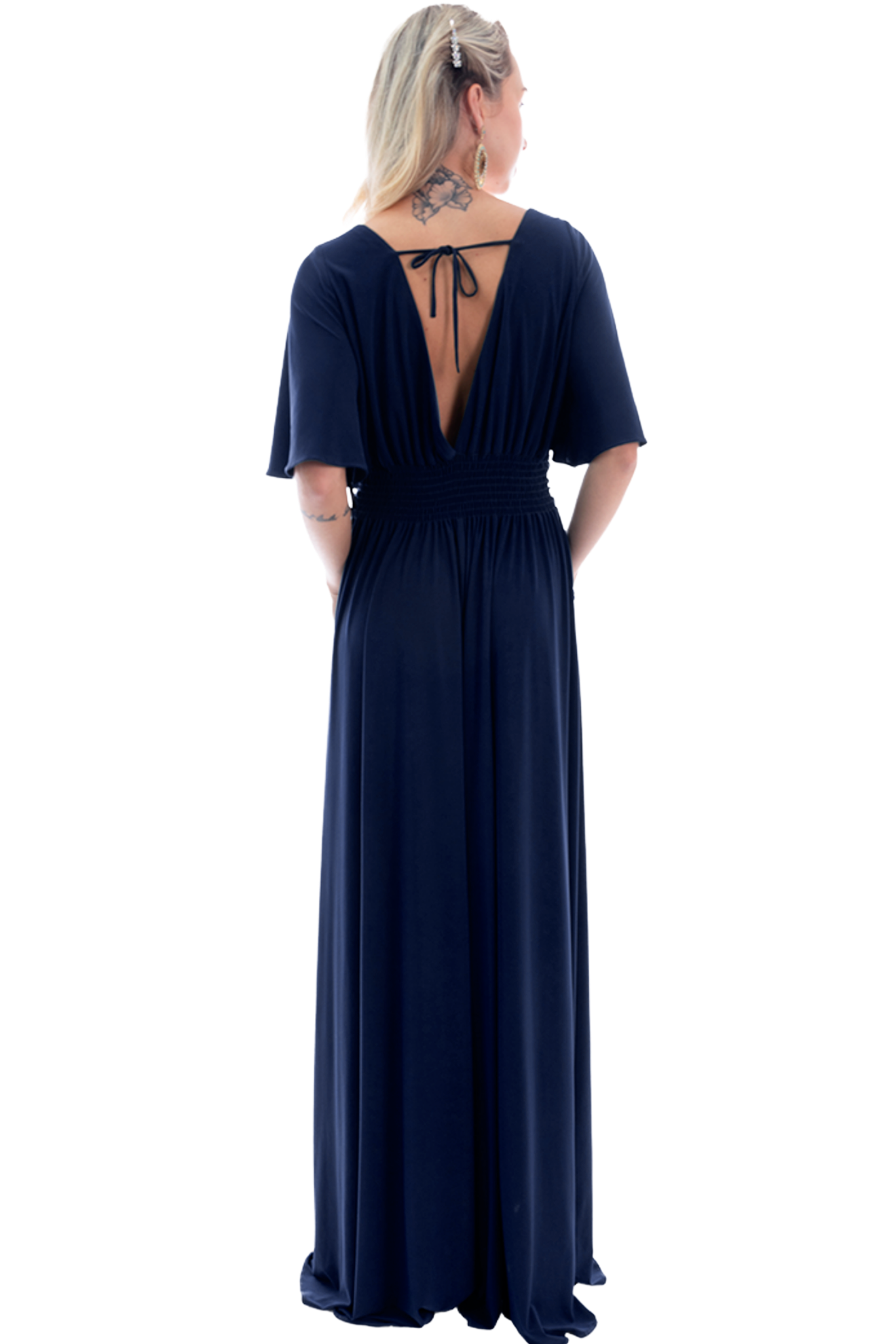 Woman seen from behind in a navy blue, floor-length dress with a deep V back and smocked waist; blonde hair with a clip and a tattoo at the neck/upper back.