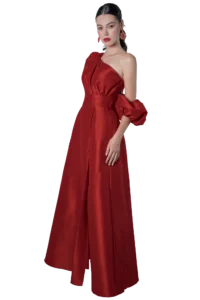 Woman in a floor-length red satin gown with one shoulder and a ruffled puff sleeve, standing against a black background wearing red lipstick and statement earrings.