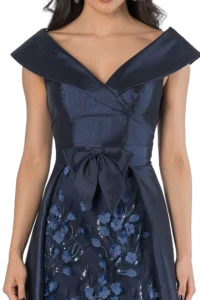 Close-up of a woman in a navy blue satin gown with a dramatic off-shoulder collar and floral-embroidered skirt piece near the waistbow area.