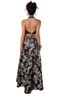 Back view of a woman wearing a black and beige floral maxi dress with a halter neck, standing against a black background