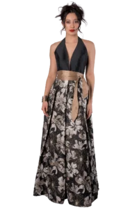 Woman in a halter black top with a bronze sash tied at the waist, and a long, floral-patterned skirt in neutral tones, standing against a black background.