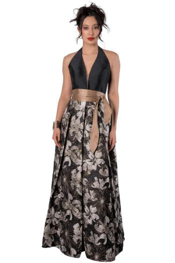 Woman in a halter black top with a bronze sash tied at the waist, and a long, floral-patterned skirt in neutral tones, standing against a black background.