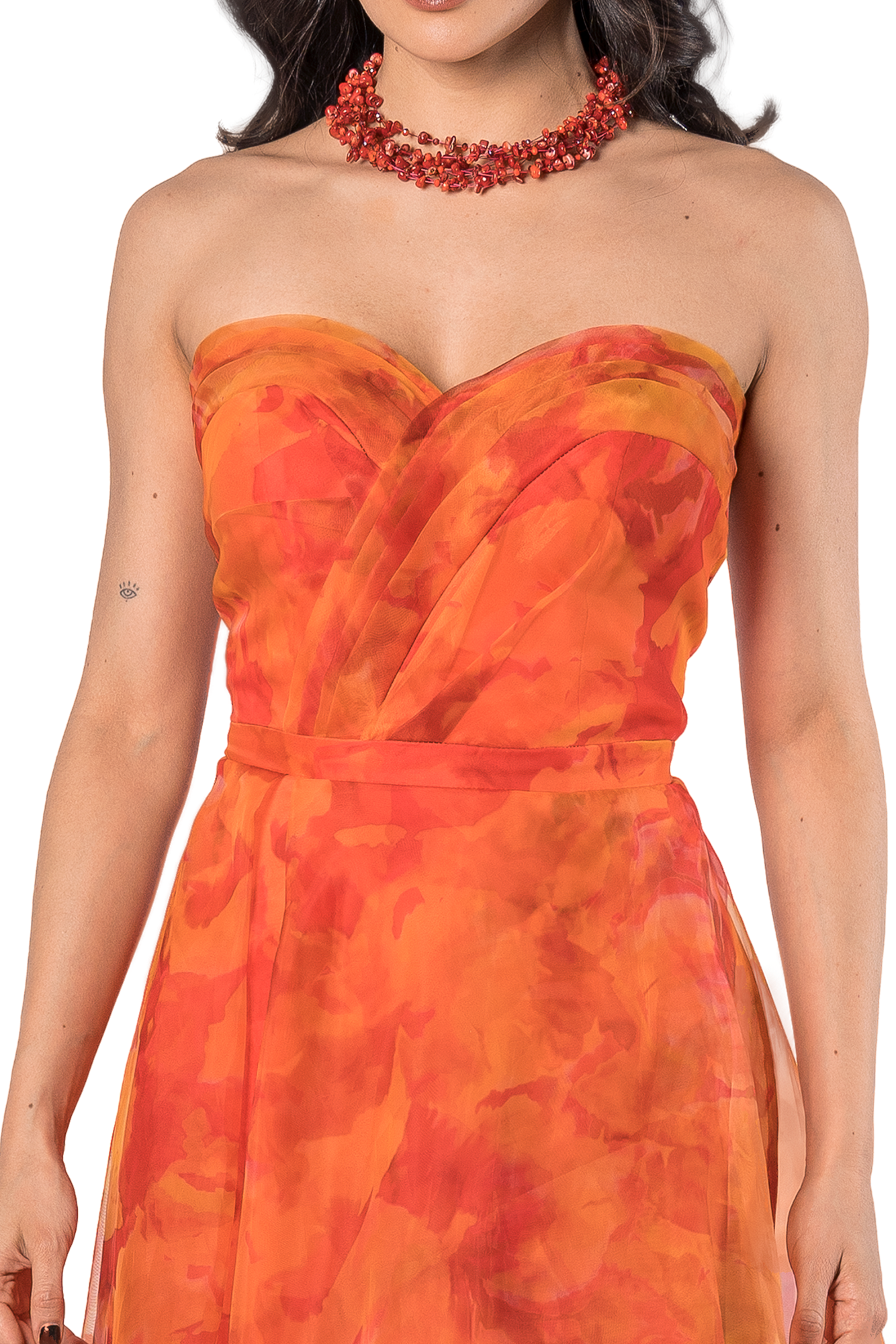 Woman in an orange strapless dress with a red beaded necklace, showing her shoulders and a small eye tattoo on her left forearm.
