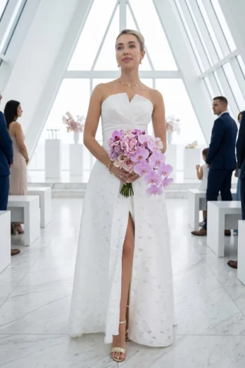 Bride in a white strapless wedding gown holding a pink orchid bouquet in a bright, modern venue.