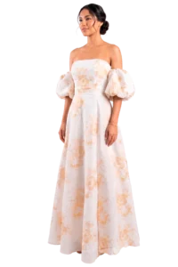 Woman in a strapless, floor-length white gown with peach floral print and puff sleeves, standing against a black background.