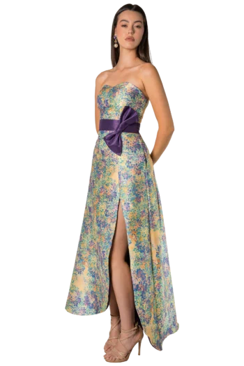 Woman in a colorful floral strapless gown with a dark purple sash bow and slit, posing against a black background.