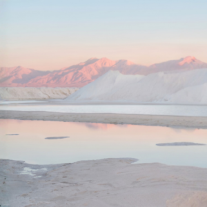 Pastel-colored mountain range at sunset over a flat salt landscape with a pale blue sky.
