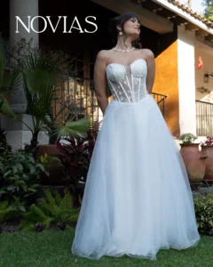 Bride wearing a white strapless corset wedding gown with a full tulle skirt, standing in a garden with the 'NOVIAS' logo in the top left corner.